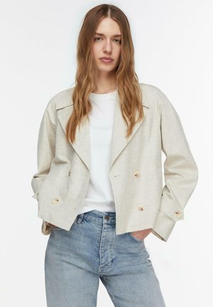 Młoda kobieta z długimi włosami, ubrana w beżową krótką kurtkę na białej koszuli i jasnoniebieskich dżinsach, stoi z rękami w kieszeniach.