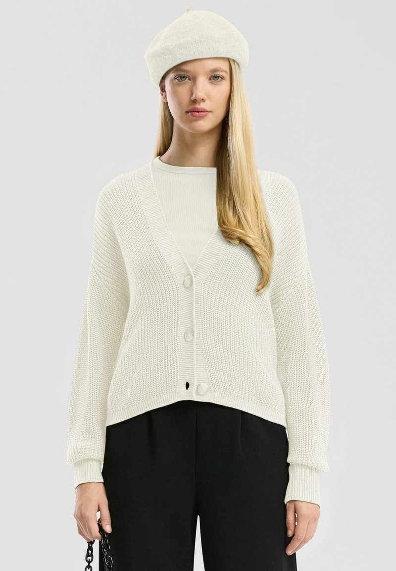 Witte, gebreide cardigan met een diepe V-hals, drie knopen en geribbelde manchetten, gecombineerd met een lichte top en zwarte broek. Minimalistisch ontwerp.