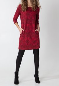 Czerwona sukienka w wzory z rękawami do łokcia, dwiema bocznymi kieszeniami i luźnym krojem, zestawiona z czarnymi rajstopami i botkami za kostkę.