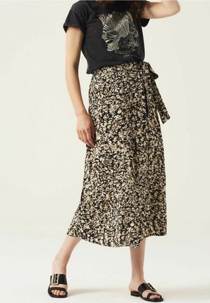 Femme portant un t-shirt noir à motifs et une jupe midi fleurie taille haute avec une ceinture nouée sur le côté, ainsi que des sandales noires à enfiler.