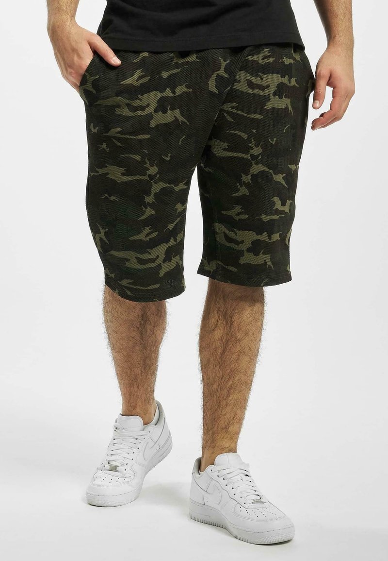 Homme portant un short camouflage vert foncé et noir, chemise noire et baskets blanches, debout les mains partiellement dans les poches.