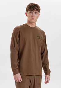 Camicia marrone a maniche lunghe in cotone, caratterizzata da un collo tondo e una grafica frontale con l'anno "1935" in stampa scura. Design semplice, vestibilità comoda.