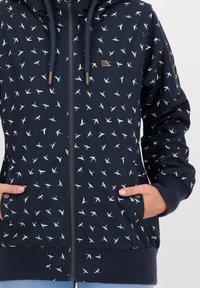 Marinblå zip-jacka med vita fågelmönster, dragsnören, ribbade muddar och nederkant, framfickor och en liten metalltagg med "WE ARE ALIFE".