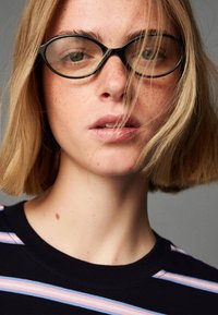 Kerek fekete szemüveg keretez egy arcot, amelyen világos szeplők láthatók, rövid szőke hajjal és egy sötétkék inget visel, amelyen vízszintes pasztell csíkok találhatók.