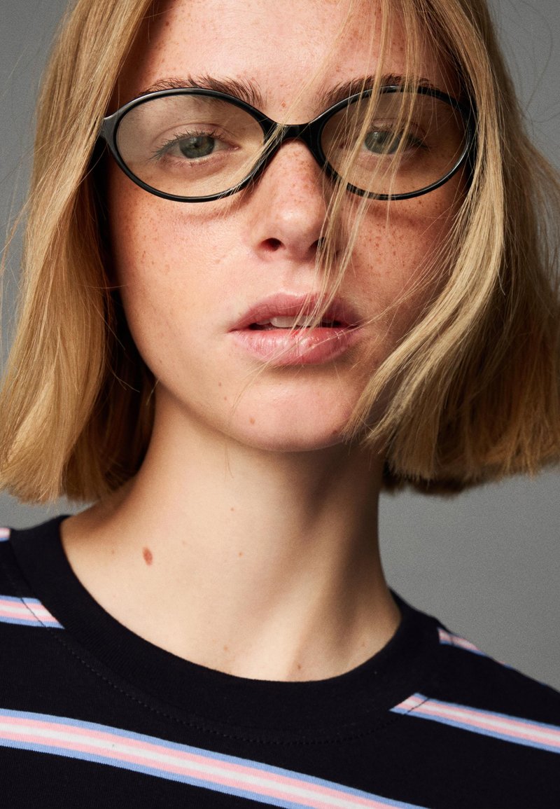 Kerek fekete szemüveg keretez egy arcot, amelyen világos szeplők láthatók, rövid szőke hajjal és egy sötétkék inget visel, amelyen vízszintes pasztell csíkok találhatók.