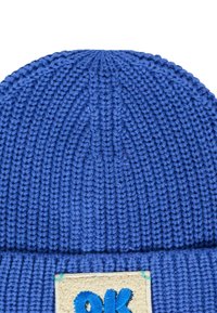 Blauwe gebreide beanie met een textuurpatroon, voorzien van een opgevouwen manchet en een rechthoekige patch met "ok" in blauwe borduuring op een crèmekleurige achtergrond.