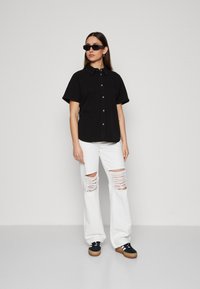 Maglietta nera a maniche corte con chiusura a bottoni, abbinata a jeans bianchi a gamba larga strappati e sneakers nere con suola in legno.