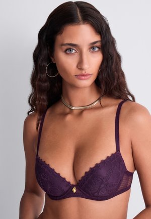 Femme aux longs cheveux ondulés et foncés porte des boucles d'oreilles créoles dorées, un collier ras-du-cou doré et un soutien-gorge en dentelle violet, posant devant un fond neutre.