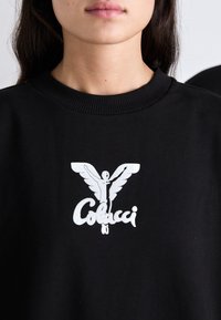 Černá mikina s žebrovaným límcem, která má velký bílý grafický motiv křídlové postavy a text "Colucci" pod ním.