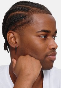 Orecchino a cerchio nero indossato all'orecchio sinistro di una persona con capelli intrecciati. L'individuo tiene il mento appoggiato su una mano, indossando una camicia bianca.