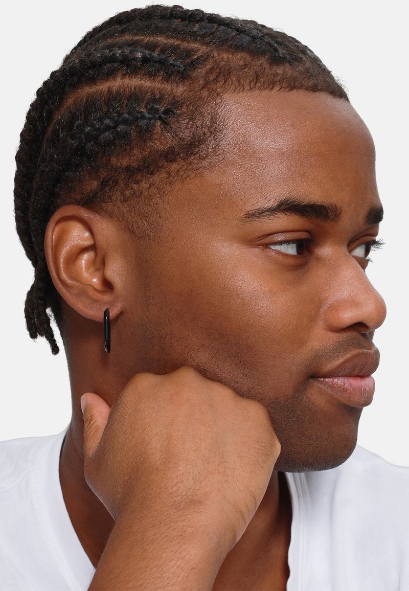 Orecchino a cerchio nero indossato all'orecchio sinistro di una persona con capelli intrecciati. L'individuo tiene il mento appoggiato su una mano, indossando una camicia bianca.