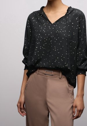 Zwarte blouse met lange mouwen en witte stippen, ingestopt in beige, hooggetailleerde broek met een rechthoekige gesp.