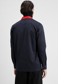 Marinblus med röd krage, långa ärmar och knappade manschetter. Har en slät textur, ryggsöm och minimalistisk design.