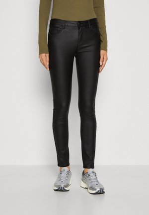 Femme portant un pantalon skinny en cuir noir, un haut à manches longues vert olive et des chaussures de sport argentées, debout devant un fond blanc.