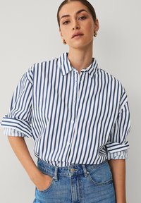 Stribet skjorte med lodrette navy og hvidstriber, en klassisk krave og opkrøllede ærmer. Parret med lyseblå jeans.