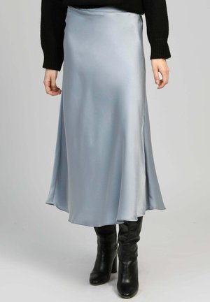 Jupe midi en satin bleu clair portée avec un pull noir et des bottes en cuir noires à talons montant jusqu'au genou, posant devant un fond neutre.