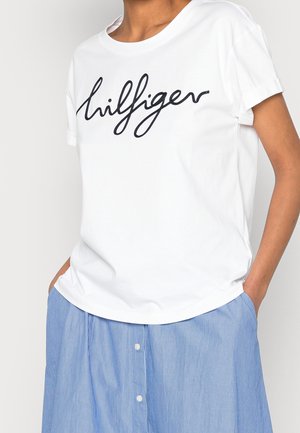 Osoba w białej koszuli z krótkim rękawem z napisem "hilfiger" oraz w niebiesko-białej paskowanej spódnicy zapinanej na guziki, z rękami w kieszeniach.