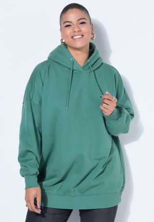 Uśmiechnięta osoba z krótkimi włosami, ubrana w za dużą zieloną bluzę z kapturem, złote kolczyki koła i ciemne spodnie na białym tle.