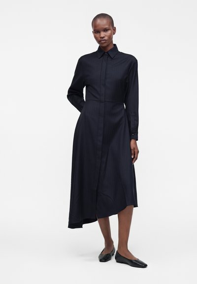 Femme grande aux cheveux courts portant une robe chemise midi noire à manches longues et des ballerines noires à bout carré, debout devant un fond blanc uni.