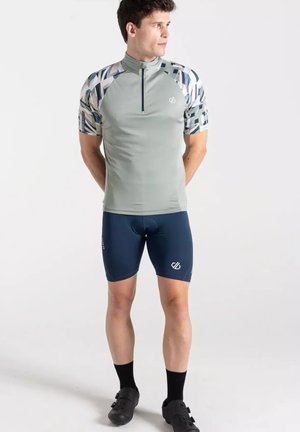 Man draagt een grijs fietsshirt met patroon op de mouwen, marineblauwe gewatteerde fietsbroek, zwarte sokken en zwarte fietsschoenen, staand met de handen achter de rug.