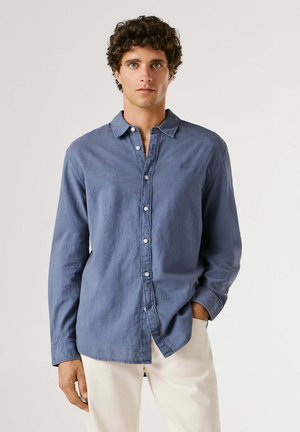 Junger Mann mit lockigem Haar, der ein blaues Hemd mit Knöpfen und hellbeige Hose trägt, steht mit einer Hand in der Hosentasche vor einem einfarbigen Hintergrund.