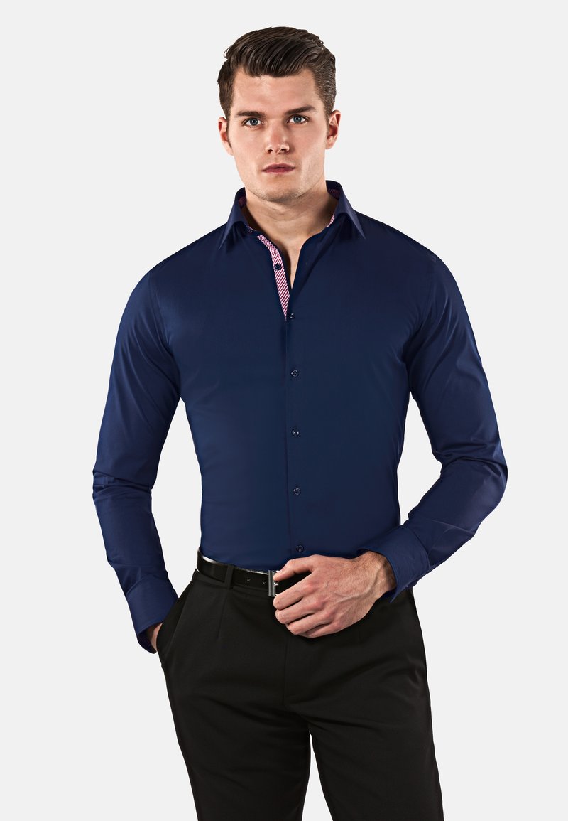 Camicia a maniche lunghe blu navy con colletto interno a quadretti in contrasto, dotata di chiusura a bottoni e una consistenza del tessuto liscia. Abbinata a pantaloni neri.