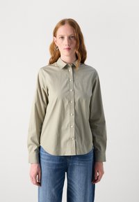 Light olive green button-up shirt with a classic collar, long sleeves, and subtle sheen; paired with blue straight-leg jeans.
