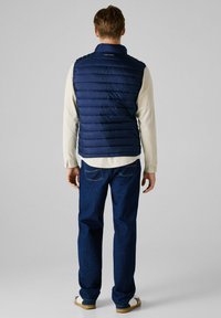 Marineblauwe gewatteerde vest met horizontale naden, hoge kraag en 'Pepe Jeans' logo, gedragen over een lichtgekleurde shirt en blauwe jeans.