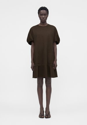 Femme aux cheveux courts portant une robe ample marron foncé, longueur genou, avec des manches courtes bouffantes et des talons ouverts noirs, debout sur un fond blanc.