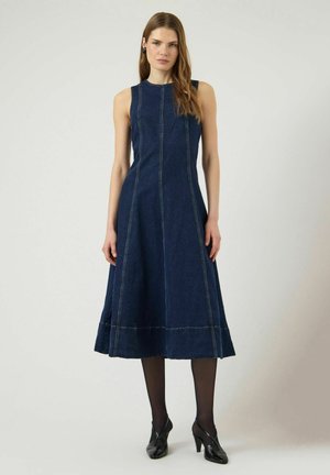 Naine, kes kannab varrukateta tumeda teksakleit midi pikkuses, musti sukkpükse ja musta kontsaga jalanõusid, seisab ühevärvilise heleda tausta ees.