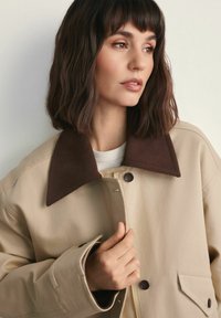 Kobieta z włosami do ramion w ciemnym kolorze, ubrana w beżową kurtkę z dużym ciemnobrązowym kołnierzem, trzymając kurtkę przy guzikach, patrzy w prawo.