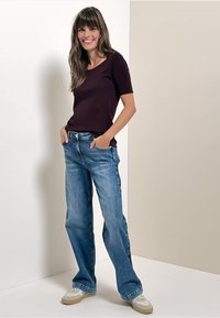 Bordeaux kortom shirt gecombineerd met lichtblauwe wijde jeans en beige sneakers, staande tegen een neutrale achtergrond.