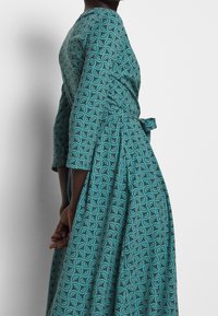 Robe bleu sarcelle à manches trois-quarts, arborant un motif tourbillonnant. Le tissu semble texturé, avec un détail de taille nouée. Vue de profil.
