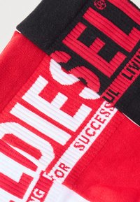 Røde og hvite strikkede sokker med svart elastisk mansjett. Har fet "DIESEL"-merking i kontrasterende farger. Teksturert stoff.