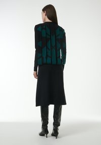 Schwarzes Kleid mit einem geometrischen Muster in Grün und Schwarz. Hat lange Ärmel, eine figurbetonte Silhouette und knielange glänzende schwarze Stiefel.