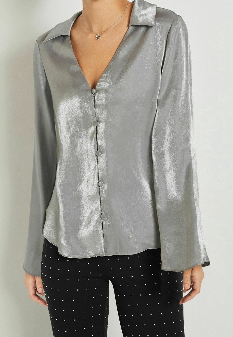 Blouse brillante argentée à manches longues boutonnée avec un col, portée avec un pantalon noir à petits pois blancs.