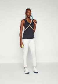 Αθλητικό τοπ σε μαύρο με λουριά σε ταμπά χρώμα, σε συνδυασμό με λευκό εφαρμοστό παντελόνι και λευκά αθλητικά παπούτσια. Κρατά ρακέτα τένις και μπάλα.