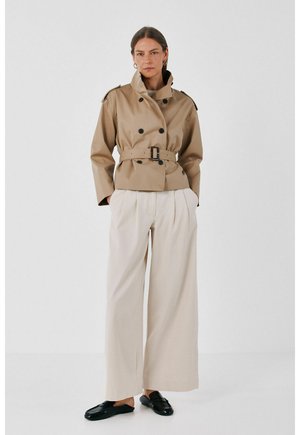 Femme portant une veste beige ceinturée à double boutonnage, un pantalon crème à jambes larges, des chaussures noires sans lacets, debout sur un fond blanc uni.