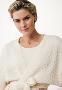 Krämfärgad, texturerad kofta med diamantmönster, knytet i midjan. Bärs över en ljusfärgad topp och accessoriserad med ett delikat halsband.