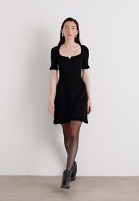 A woman walks confidently in a black short dress with puff sleeves, paired with black ankle boots, against a neutral background.
