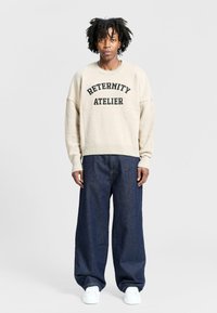 Beige knit sweatshirt with "ETERNITY ATELIER" in black text, paired with wide-leg dark denim jeans and white sneakers.