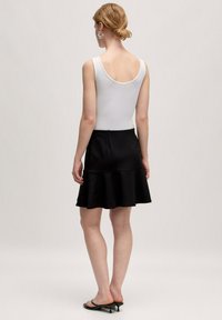 Weißes Tanktop und schwarzer ausgestellter Rock, der eine glatte Textur zeigt. Der Rock hat ein schichtweises Design und sitzt oberhalb des Knies.