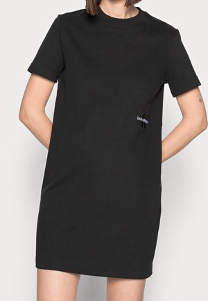 Femme portant une robe courte noire décontractée à manches courtes avec un petit logo Calvin Klein sur la hanche gauche.