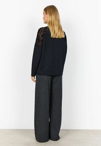 Black long-sleeve top with lace detailing on shoulders, paired with wide-leg gray trousers. Smooth fabric texture, relaxed fit design.