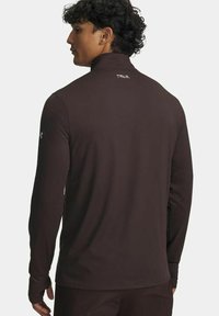 Man met krullend haar die zijwaarts kijkt, met een aansluitende zwarte sporttop met lange mouwen en duimgaten, met het "TRAIL"-logo op de bovenrug.