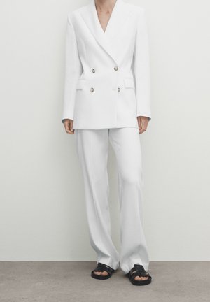 Personne portant un blazer blanc croisé et un pantalon large assorti, avec des sandales noires à ouvertures sur les orteils, posant devant un mur uni.