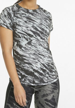Mujer con camiseta deportiva de manga corta, ajustada, con estampado en gris y negro y mallas a juego sobre un fondo blanco liso.