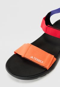 Les sandales Terrex présentent une semelle noire, des sangles ajustables orange et rouges, ainsi qu'un accent violet. Les sangles texturées arborent un détail de logo.
