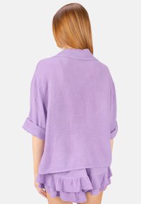 Lavendelfarbene, strukturiert gestrickte Bluse mit umgeschlagenen Ärmeln und lockerer Passform, kombiniert mit passenden, rüschengeschmückten Shorts. Rückansicht gezeigt.
