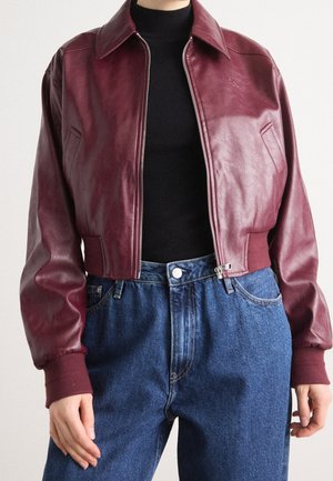 Chaqueta bomber de cuero rojo oscuro recortada, usada sobre un jersey de cuello alto negro y vaqueros azules de talle alto con botones y cremallera en la parte delantera.